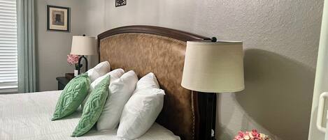 A neatly made bed with white and green pillows, a wooden headboard, a bedside lamp, and a small table with a vase of flowers.