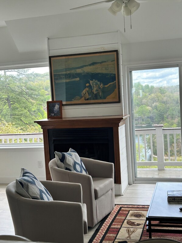 A living room with a fireplace, a painting above it, and a view of a forested area through large windows.