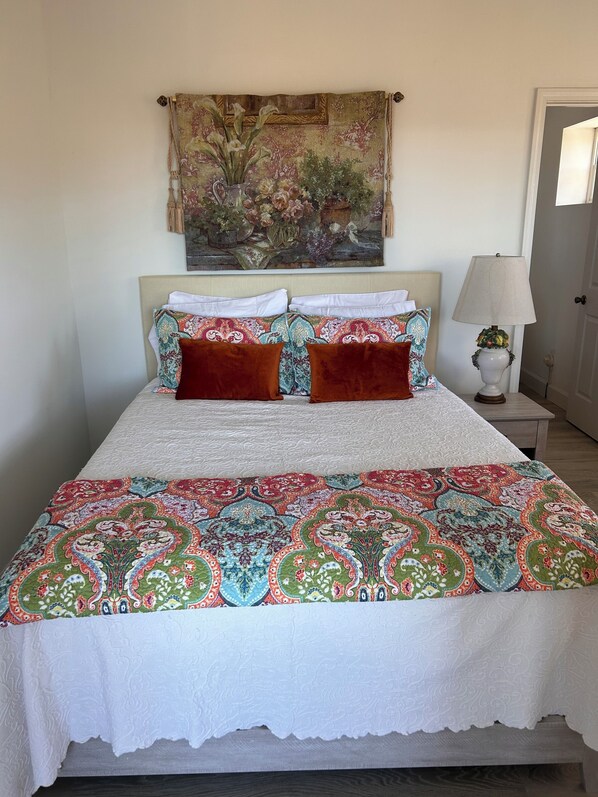 Una cama con una colcha estampada, dos almohadas, una mesita de noche con una lámpara y un tapiz en la pared.