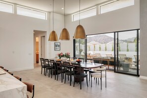 A spacious dining area with a large table and chairs, a modern pendant light fixture, and a view of the outdoors through large glass doors. - The Reserve at Polo Villas by AvantStay (La Quinta)