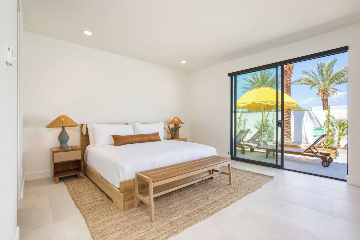 A bedroom with a large bed, two bedside tables, a bench, and a view of palm trees through a sliding glass door.