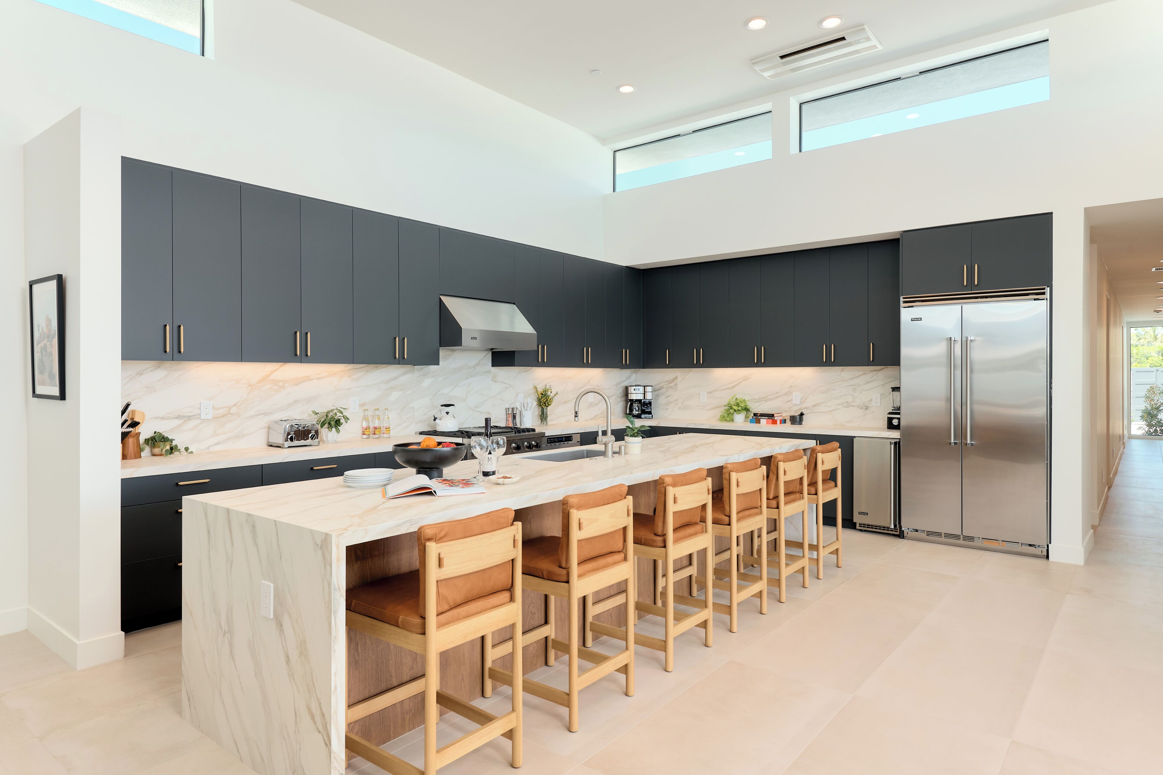 A modern kitchen with dark cabinets, a large island with bar stools, a stainless steel refrigerator, and a skylight.