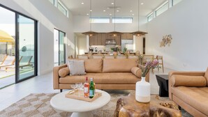 A spacious living area with a large sofa, a coffee table, and a dining table. There is a kitchen in the background with modern appliances and a dining area with chairs. - The Reserve at Polo Villas by AvantStay (La Quinta)