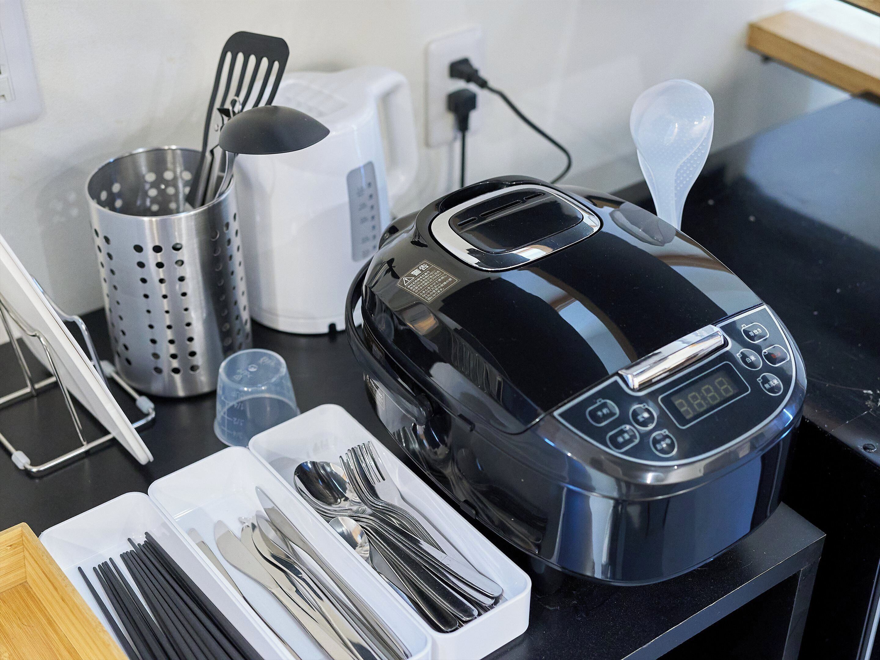 A kitchen counter with a rice cooker, a colander, a toaster, and a set of utensils.