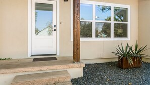 A house with a white door, a window with a view of trees, and a potted plant on the porch. - The Corral | 2/1, Hot Tub, Full Kitchen, Gas Grill, Gazebo (Fredericksburg)