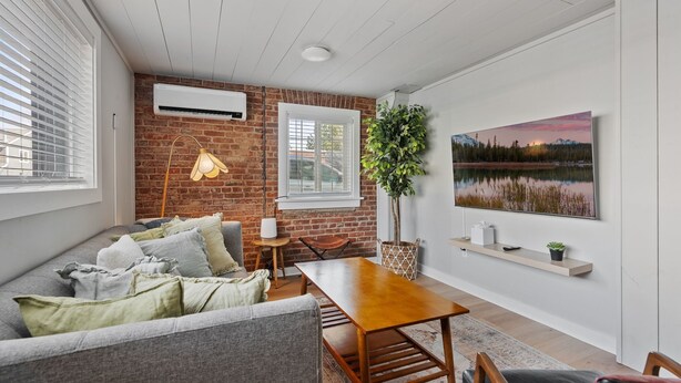 A cozy living room with a brick wall, a wooden table, a sofa with cushions, a potted plant, and a painting on the wall. - Stylish Downtown Cottage, Walk Everywhere (Bend)