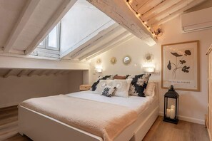 A bedroom with a skylight, a bed with floral bedding, wall sconces, and a framed botanical print.