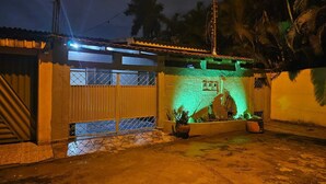 Un edificio con entrada protegida por una reja, un pequeño jardín con plantas en macetas y un cuadro eléctrico a la vista en la pared.