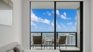 Et moderne soveværelse med en stor seng, udsigt over byen gennem gulv til loft-vinduer, og en balkon med stole.