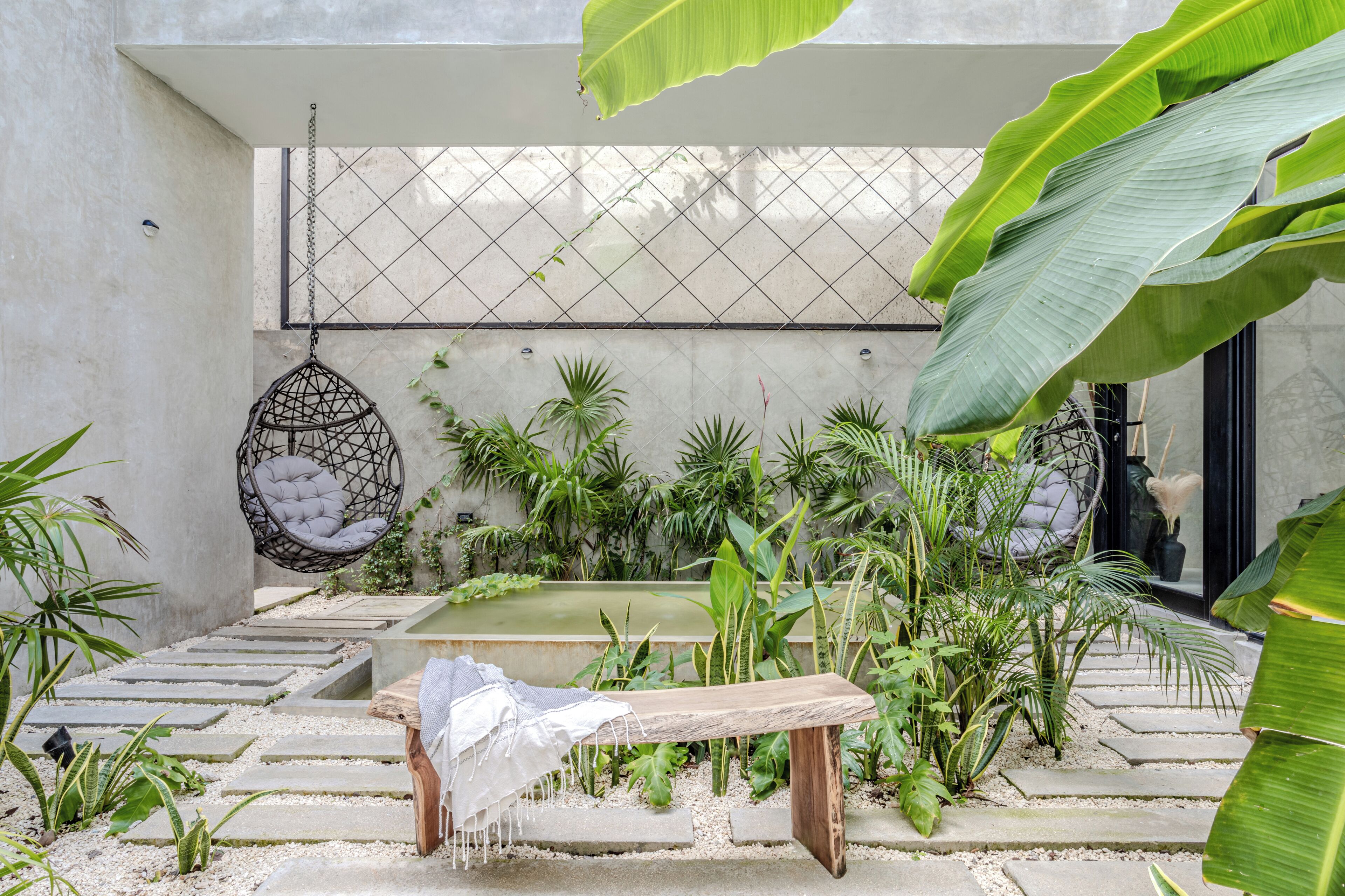 Un jardin paisible avec un banc en pierre, un fauteuil suspendu et diverses plantes.
