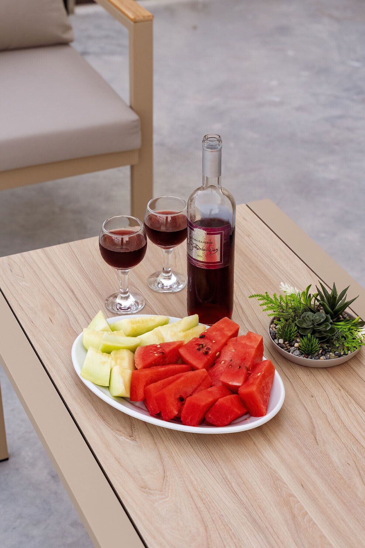 A wooden table with a plate of watermelon and melon slices, a bottle of red wine, and two wine glasses.