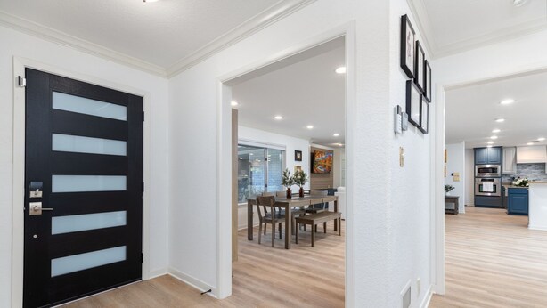 A modern interior with a black door, wooden flooring, a dining area with a table and chairs, and a kitchen in the background.