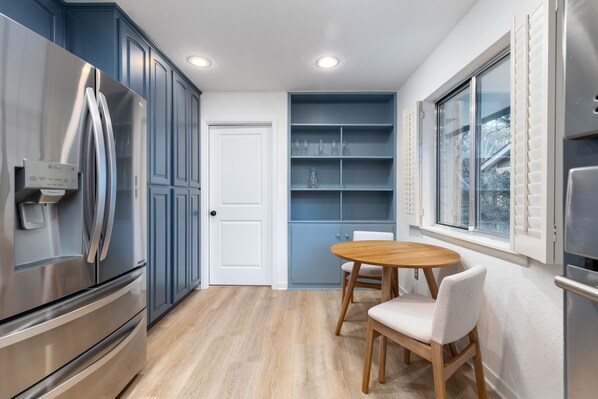 A modern kitchen with blue cabinets, a stainless steel refrigerator, a wooden dining table with two chairs, and a window with blinds. - Centrally located cozy home close to everything. (San Antonio)