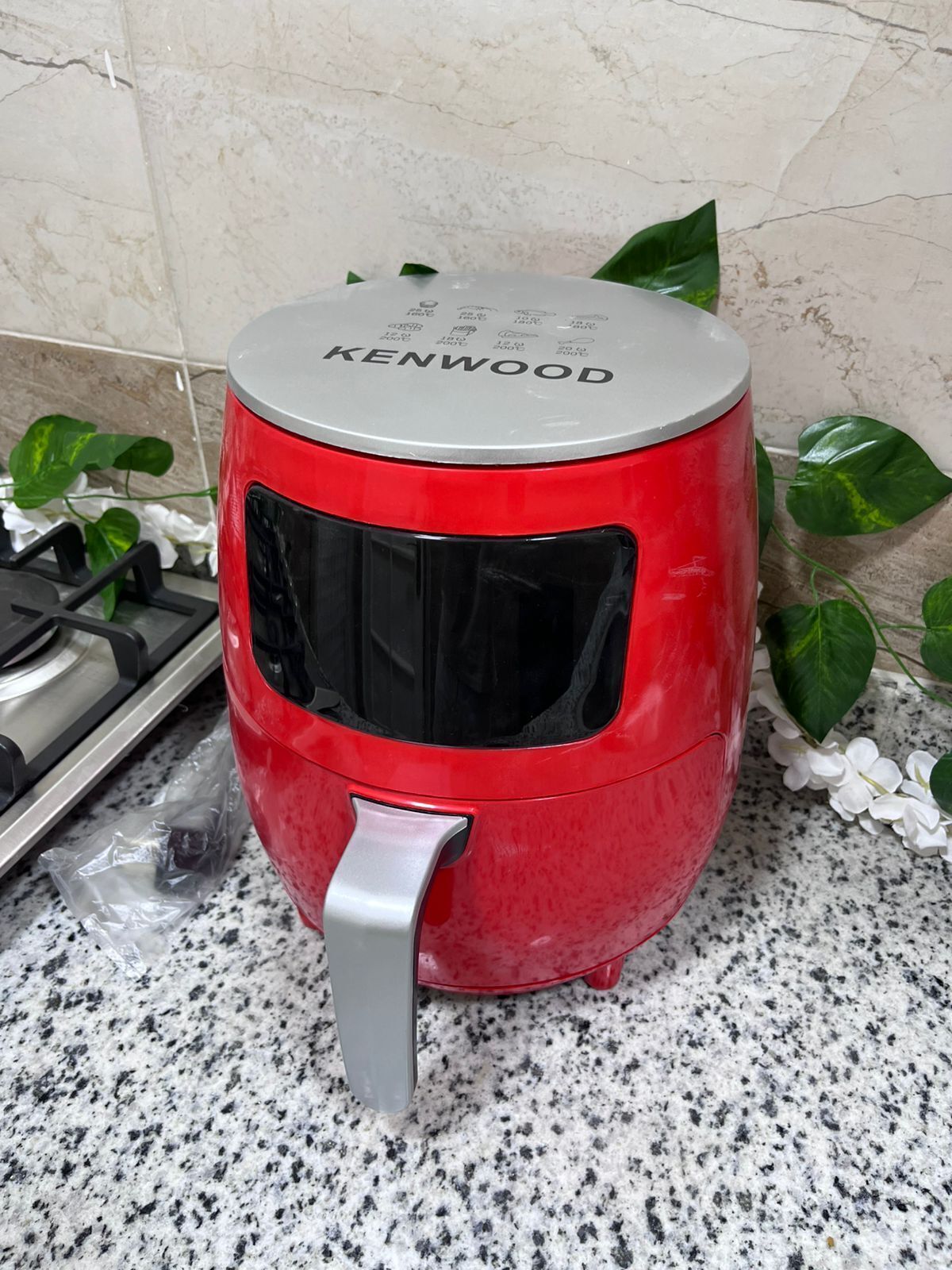 Une friteuse à air rouge de la marque Kenwood, posée sur un plan de travail en granit, avec une cuisinière en acier inoxydable et un sac en plastique à proximité.