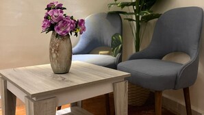 Une petite table en bois sur laquelle est posé un vase rempli de fleurs violettes, un fauteuil gris et une grande plante verte dans un coin d’une pièce dont le sol est recouvert de parquet.