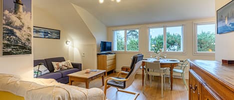 A spacious living area with a sofa, coffee table, and dining table. There is a TV on a stand and a painting of a lighthouse.