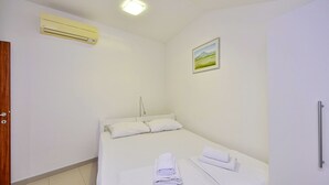 A small, clean room with a bed, a bedside table, a wall-mounted air conditioner, and a painting.