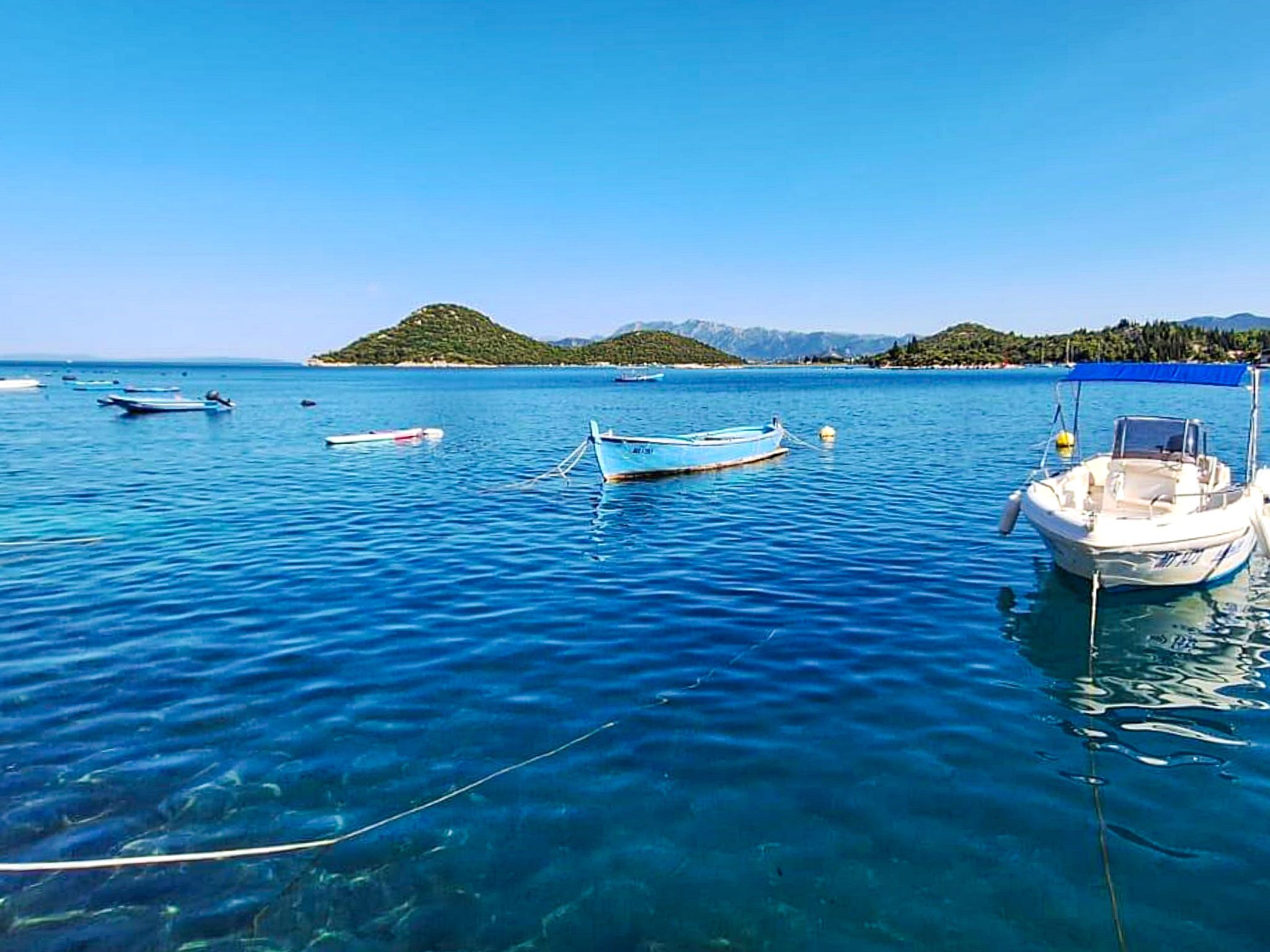 Ruhige See mit mehreren vor Anker liegenden Booten, klarer Himmel und hügelige Landschaft im Hintergrund.