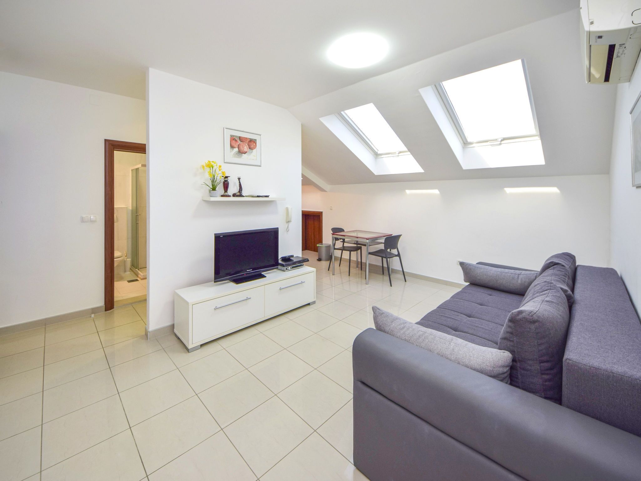 Una sala de estar moderna con un sofá gris, un televisor de pantalla plana, una pequeña mesa de comedor y un tragaluz en el techo.