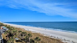 A beach with a clear sky, a sandy shore, and a few buildings in the distance.