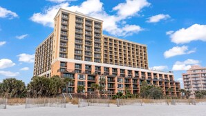 Ein mehrstöckiges Hotel mit Balkonen, gelegen an einem Strand mit Palmen und blauem Himmel.