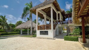 Un club de playa con un pabellón de techo de paja, una escalera que conduce a un balcón, y un cartel que indica servicios como un restaurante y una piscina.