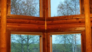 Interior de uma cabana de madeira com uma grande janela com vista para uma área arborizada e um corpo de água.