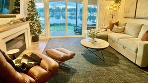 Een woonkamer met een open haard, leren fauteuils, een bank, een salontafel en uitzicht op een meer door grote glazen deuren.