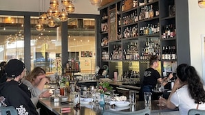 Een bar met gasten die aan tafels zitten, een barman achter de bar en kasten vol flessen.