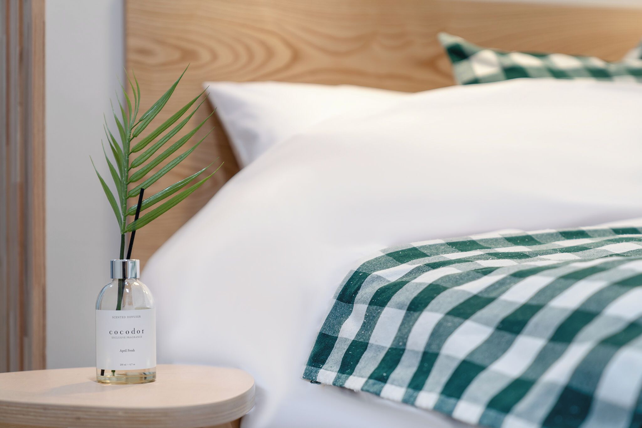 A neatly made bed with a checkered blanket, a bottle of Cocodor April Fresh on a bedside table, and a green plant.