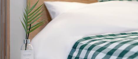 A neatly made bed with a checkered blanket, a bottle of Cocodor April Fresh on a bedside table, and a green plant.