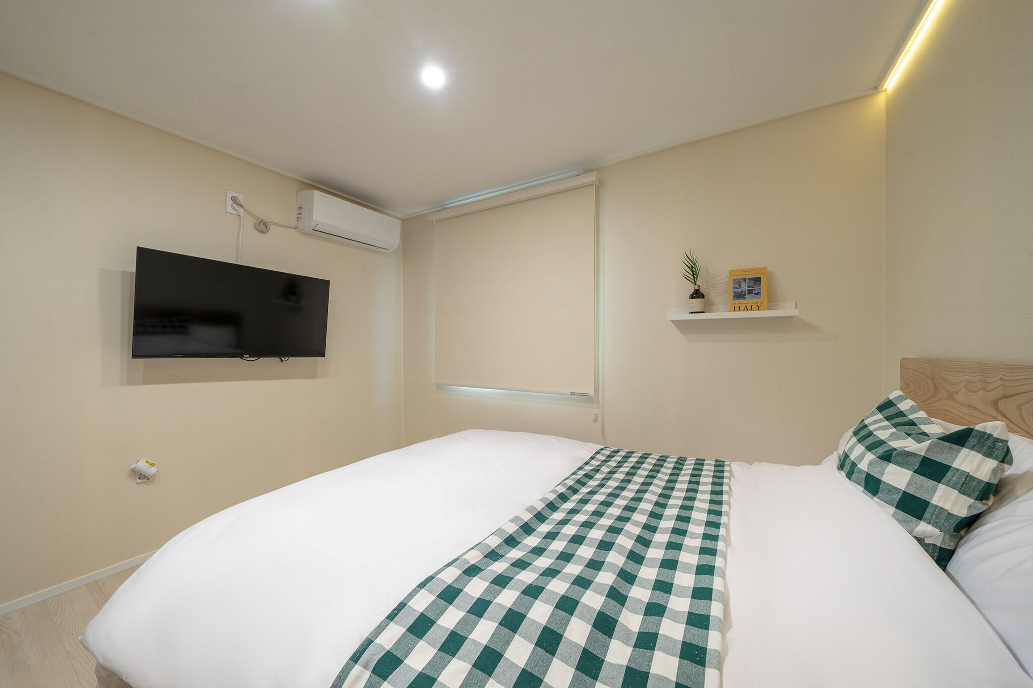 Une chambre d’hôtel avec un lit, un téléviseur fixé au mur, un appareil de climatisation, une petite étagère avec une plante et un cadre photo, et une fenêtre avec des stores.