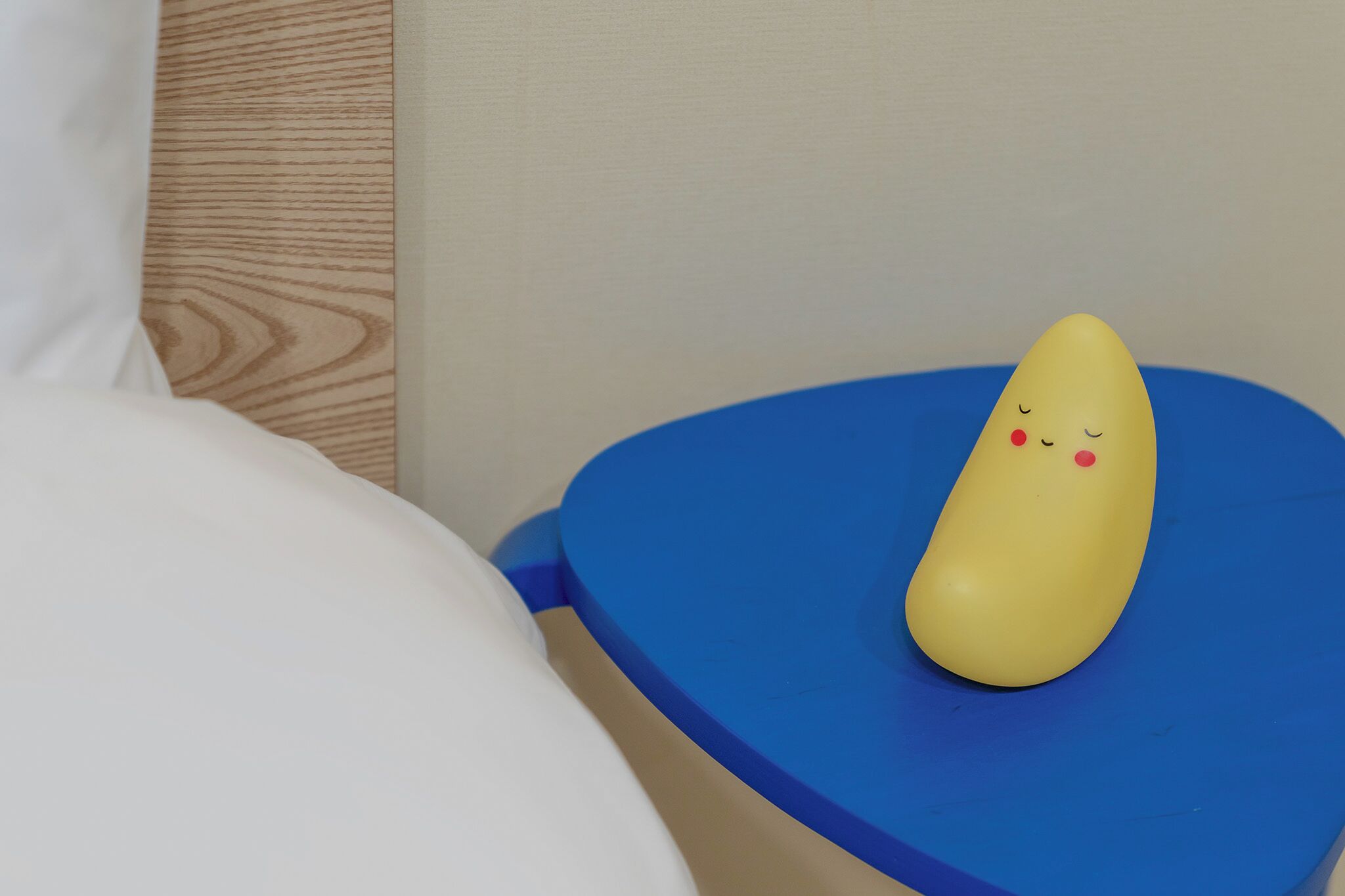 Un objeto amarillo con rostro sobre una superficie azul, junto a un almohadón blanco y un panel de madera.