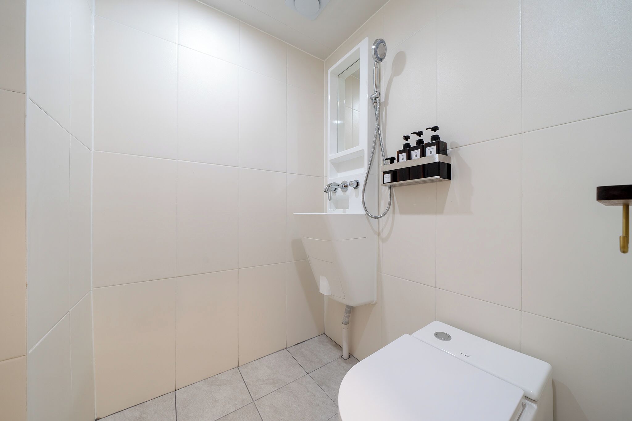A bathroom with white tiles, a shower, a toilet, and a shelf with toiletries.