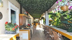 Un espacio exterior cubierto para comer, con cocina y barra, rodeado de plantas y follaje.