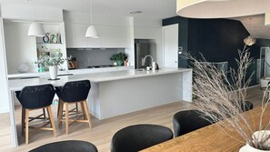 A modern kitchen with white cabinets, a central island, bar stools, and a dining area with a wooden table and black chairs. - Moonee Retreat - 200m to beach and ocean reserve views (Catherine Hill Bay)