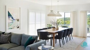 A modern living room with a sofa, a coffee table, a dining table with chairs, and a large window with blinds. - Moonee Retreat - 200m to beach and ocean reserve views (Catherine Hill Bay)
