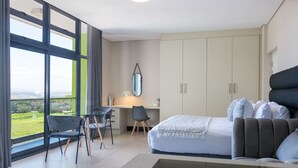 A modern bedroom with a large bed, a desk, and a chair. There is a view of greenery outside the window. - Cute and Cosy Studio Marina 501 (uMhlanga Ridge, KwaZulu-Natal)