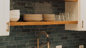 A kitchen with dark green tiled backsplash, wooden shelves, and a stainless steel sink.