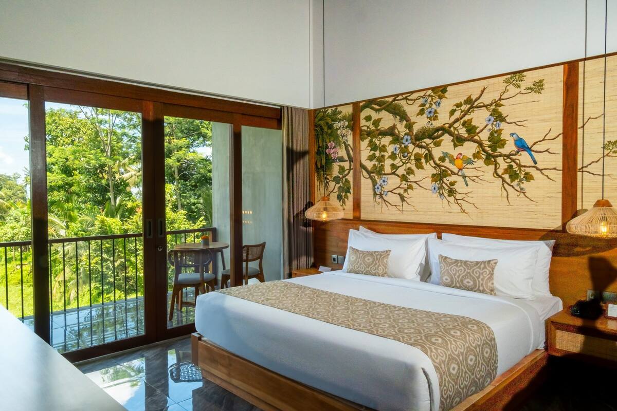 A hotel room with a large bed, a balcony with a view of greenery, and a wall mural of birds on a tree.