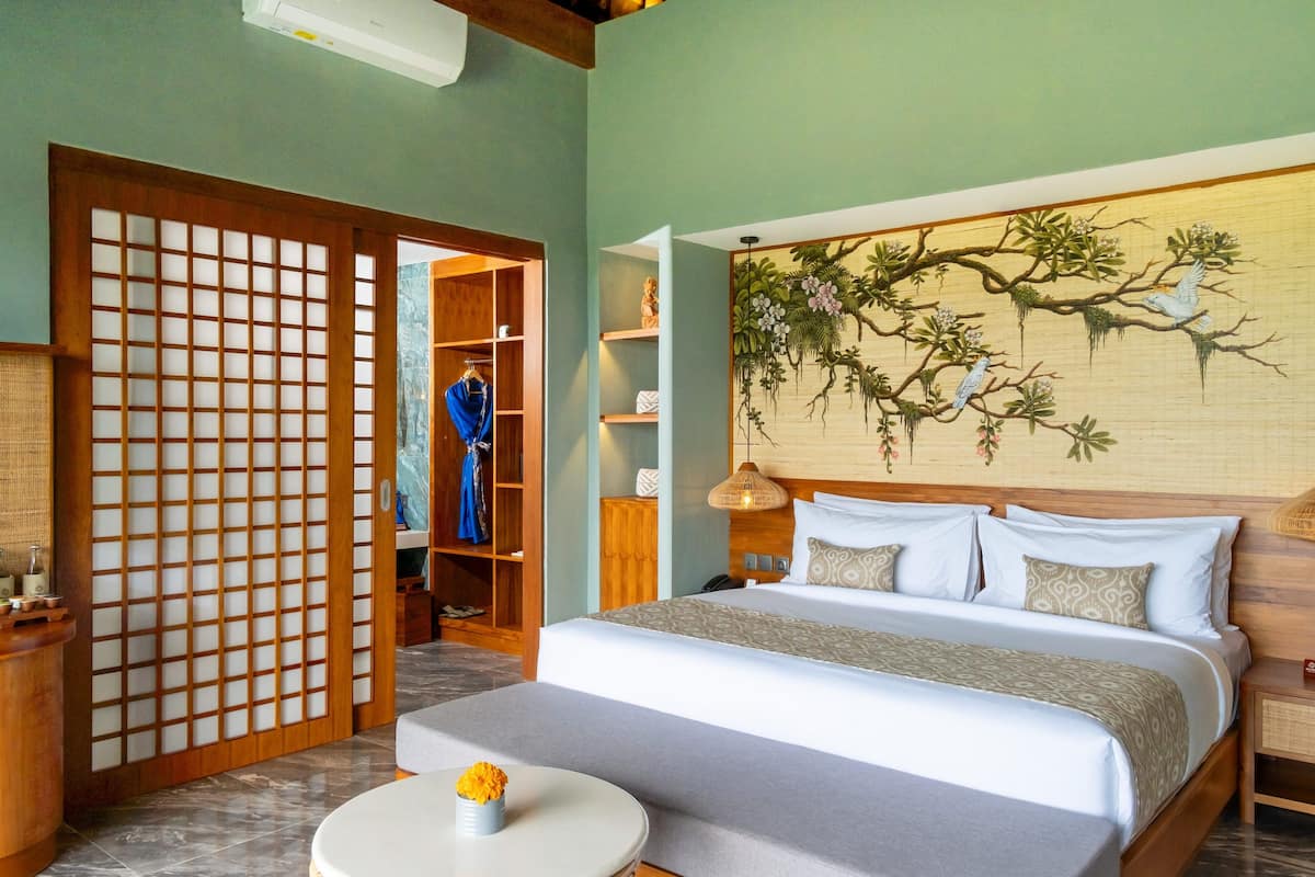 A hotel room with a bed, a small table with a vase, a wooden wardrobe, and a decorative wall painting.