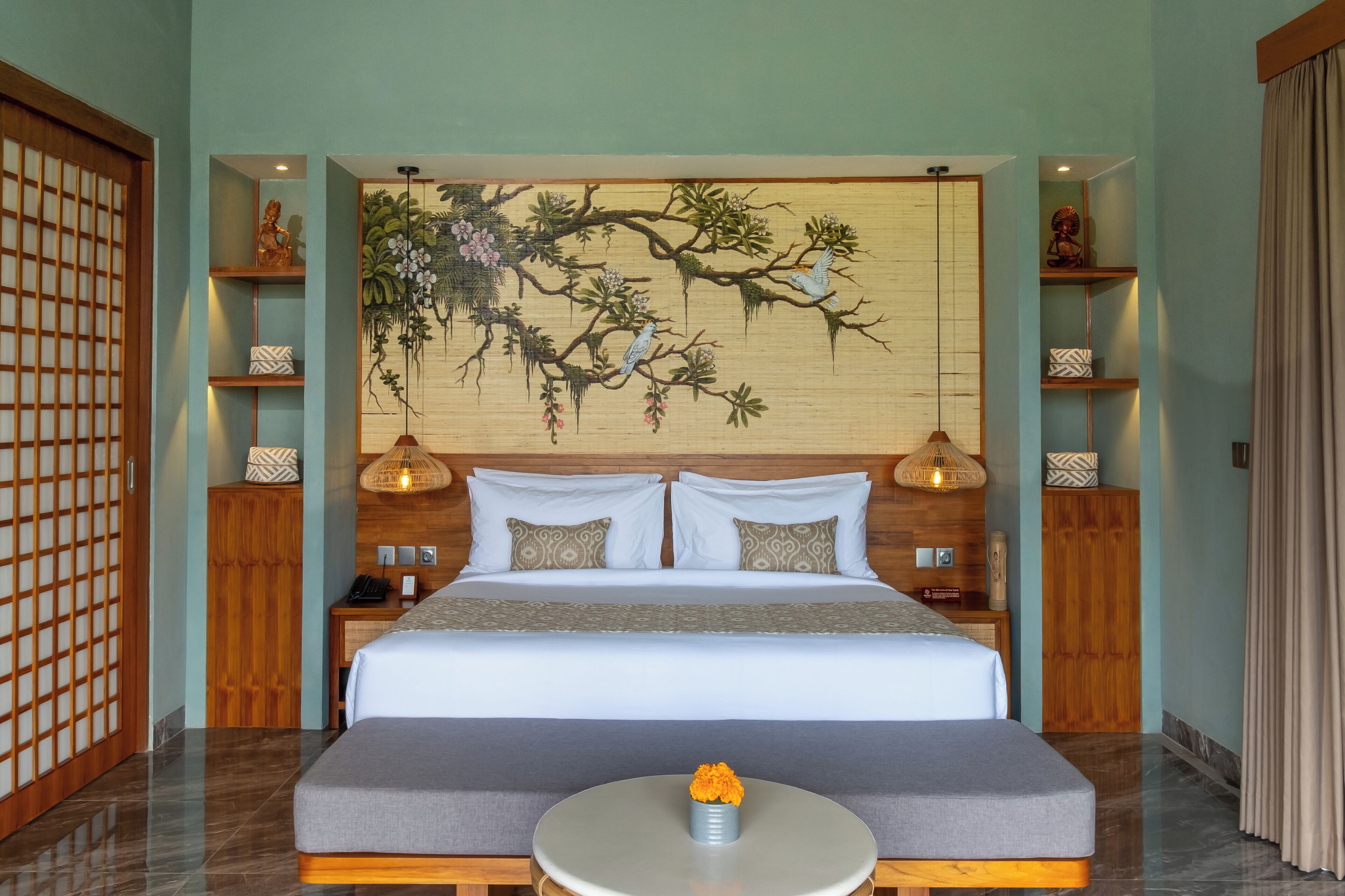 A modern hotel room with a large bed, a round table with a flower centerpiece, and a traditional-style wall hanging.