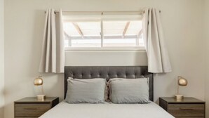 A bedroom with a tufted headboard, a large window with curtains, two bedside tables with lamps, and a bed with pillows.