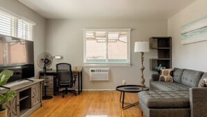 A living room with a sofa, a TV, a desk with a chair, a standing fan, and a window with blinds.