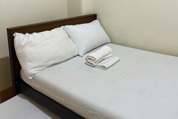Una cama bien tendida con sábanas y almohadas blancas, una toalla doblada sobre la cama y un cabecero de madera.