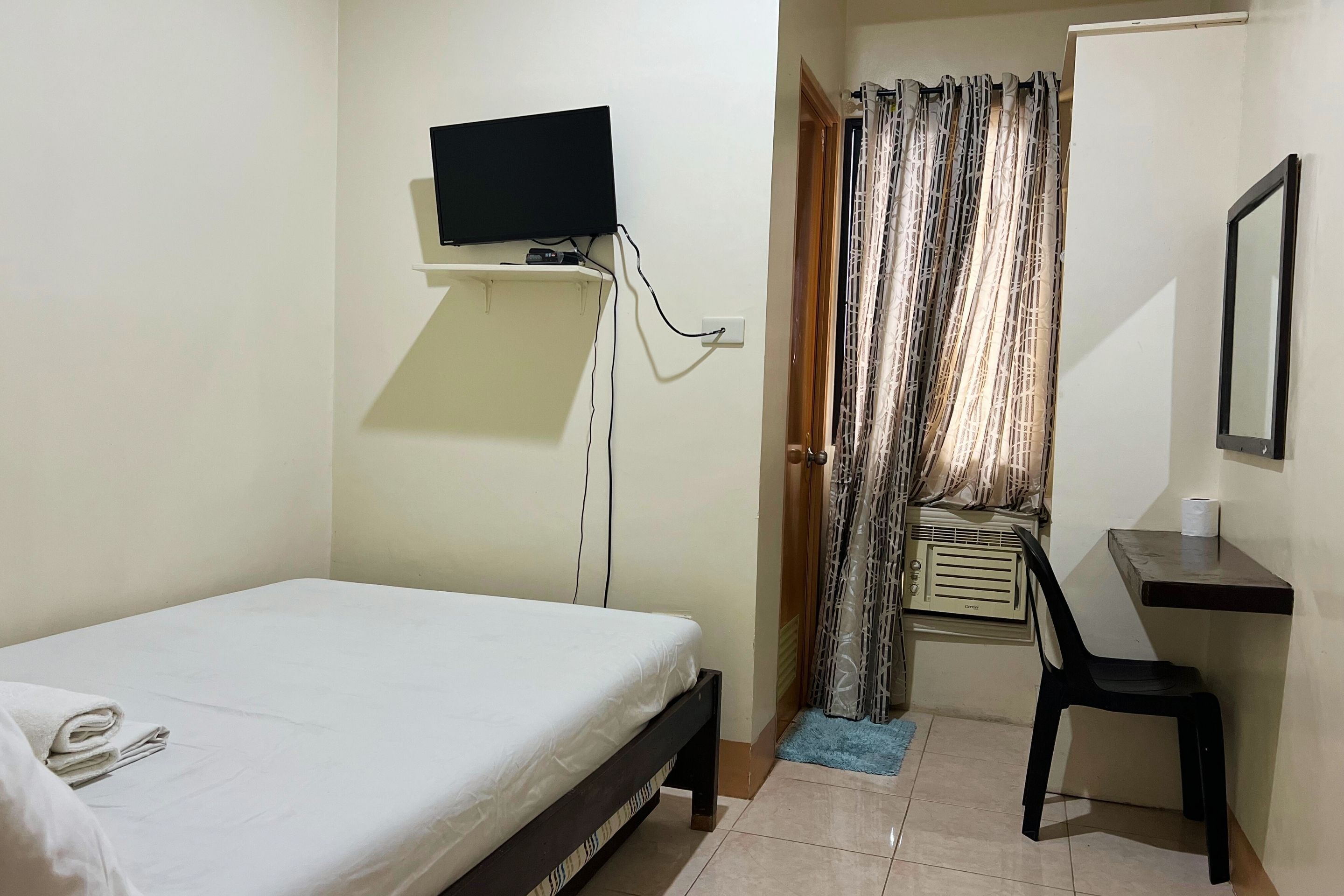 A small, clean room with a bed, a desk, a chair, a TV mounted on the wall, and a window with curtains.