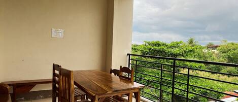 Un balcón con mesa y sillas de madera, barandilla de metal negro, y vistas a zonas verdes y edificios.