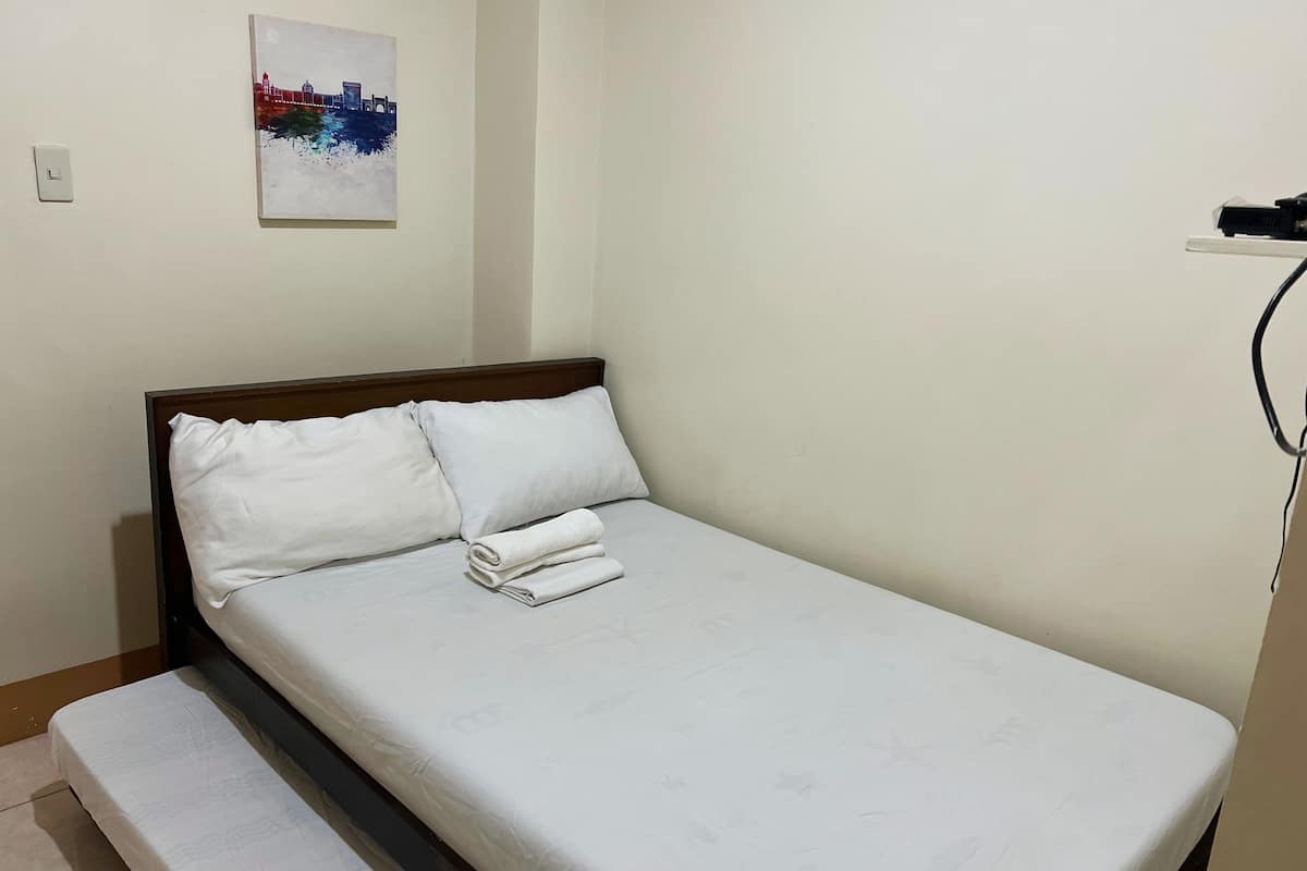 A single bed with white linens and pillows, a small bench at the foot of the bed, a wall-mounted power outlet, and a framed picture above the bed.