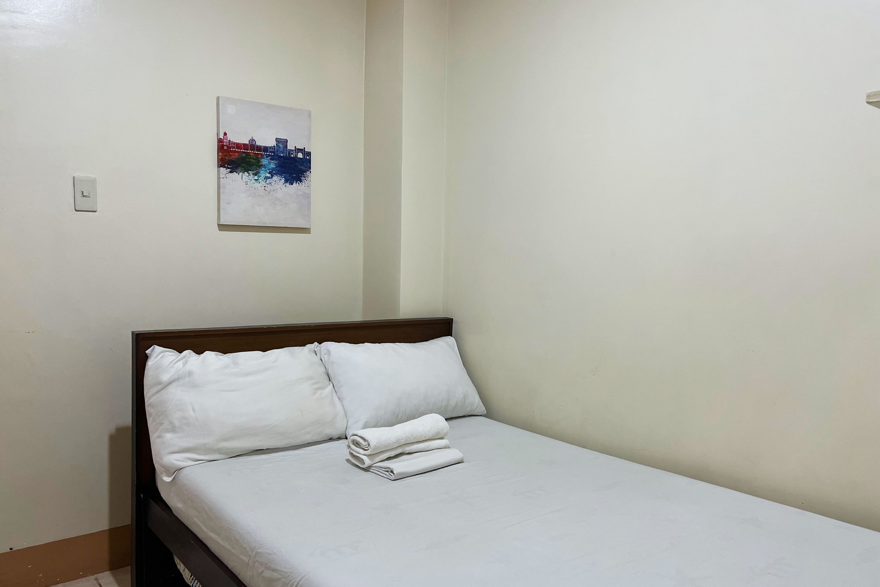 A neatly made bed with white linens and pillows in a simple room with a single wall-mounted painting.