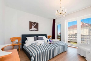 Ein Hotelzimmer mit einem Bett, einem Holzstuhl, einem kleinen Tisch, einer Chandelüster, einem Fenster mit Blick und einem Bild an der Wand.